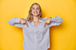 © Asier - Young blonde Caucasian woman in a striped business shirt on a yellow background, points down with fingers, positive feeling.