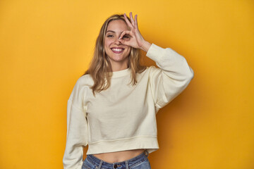 Wall Mural - Young blonde Caucasian woman in a white sweatshirt on a yellow studio background, excited keeping ok gesture on eye.