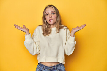 Wall Mural - Young blonde Caucasian woman in a white sweatshirt on a yellow studio background, confused and doubtful shrugging shoulders to hold a copy space.