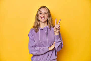 Wall Mural - Young blonde Caucasian woman in a violet sweatshirt on a yellow background, showing number two with fingers.