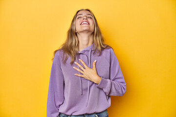 Wall Mural - Young blonde Caucasian woman in a violet sweatshirt on a yellow background, laughs out loudly keeping hand on chest.