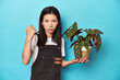 Young Asian gardener holding plant, studio backdrop, showing fist to camera, aggressive facial expression.