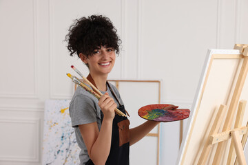 Poster - Young woman holding brushes and artist`s palette near easel with canvas