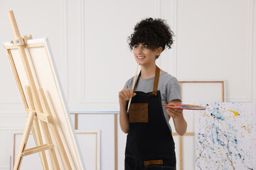 Poster - Young woman holding brush and artist`s palette near easel with canvas