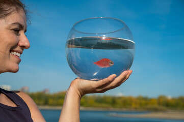 Sticker - Woman holding round aquarium with goldfish on blue sky background. 