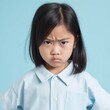 Portrait of an angry Asian little girl with black straight hair. Closeup face of a furious Chinese child on a blue background. Unhappy Japanese kid with no expression in a blue shirt looking at camera