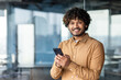 © Liubomir - Portrait of a young businessman inside the office, hispanic man is smiling and looking at the camera, the man is holding a phone in his hands, typing a message and browsing the internet.