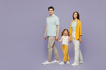 Full body side view young happy parents mom dad child kid daughter girl 6 years old wear blue yellow casual clothes hold hands walk go stroll isolated on plain purple background. Family day concept.