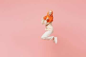 Full body young arabian asian muslim woman wears orange abaya hijab jump high do winner gesture celebrate clenching fists say yes isolated on plain pink background. Uae middle eastern islam concept.