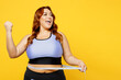 Young overweight plus size big fat fit woman wearing blue top warm up training hold measure tape on waist do winner gesture isolated on plain yellow background studio home gym. Workout sport concept.