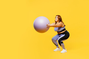 Full body side view young chubby plus size big fat fit woman wear blue top warm up training hold in hands fit ball do squats isolated on plain yellow background studio home gym. Workout sport concept.