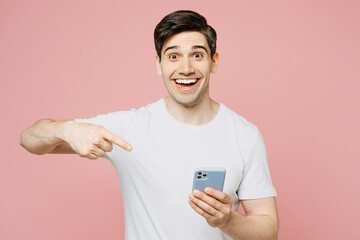 Wall Mural - Young surprised happy man wear white t-shirt casual clothes hold in hand use point index finger on mobile cell phone isolated on plain pastel light pink background studio portrait. Lifestyle concept.