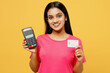Young Indian woman wearing pink t-shirt casual clothes hold wireless modern bank payment terminal to process acquire credit card isolated on plain yellow background studio portrait. Lifestyle concept.