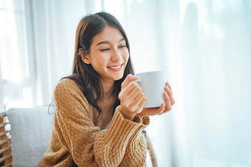 Wall Mural - Happy asian woman relaxing drinking hot coffee or tea in holiday morning vacation on armchair at home, Cosy scene, Smiling pretty woman drinking hot tea in autumn winter.