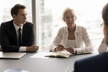 Canvas Print - Serious senior employer woman and HR recruit team talking to job candidate at interview. Different aged business coworkers s in formal clothes meeting at table, talking, discussing partnership terms