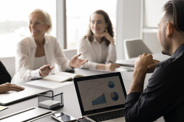 Wall Mural - Cropped shot of young business professional man speaking to happy colleagues, partners, telling marketing report on laptop, sitting at meeting table. Businessman with coworkers on blurred background