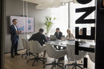 Wall Mural - Business meeting through glass wall view. Confident positive presenter man standing at electronic board in modern conference room, talking to team of colleagues, employees sitting at table