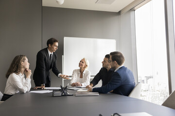 Wall Mural - Handsome young business leader man standing at meeting table in boardroom, talking to sitting listening employees. Corporate teacher training work group. Team discussing tasks, planning strategy