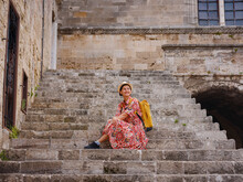 Young Asian Woman In Red Dress Walks Through Streets Of Rhodes. Woman Resting On Old House Steps In Fortress Rhodes. Tourism, Vacation, And Discovery Concept, Female Traveler Visiting Southern Europe.
