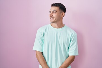 Wall Mural - Handsome hispanic man standing over pink background looking away to side with smile on face, natural expression. laughing confident.