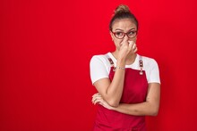 Young Hispanic Woman Wearing Waitress Apron Over Red Background Smelling Something Stinky And Disgusting, Intolerable Smell, Holding Breath With Fingers On Nose. Bad Smell