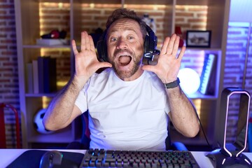 Wall Mural - Middle age man with beard playing video games wearing headphones smiling cheerful playing peek a boo with hands showing face. surprised and exited