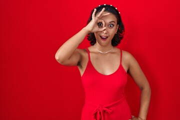 Poster - Young hispanic woman standing over red background doing ok gesture shocked with surprised face, eye looking through fingers. unbelieving expression.