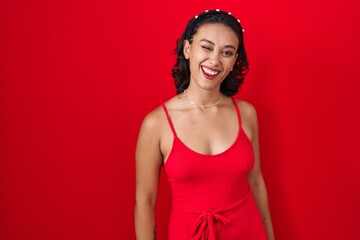 Sticker - Young hispanic woman standing over red background winking looking at the camera with sexy expression, cheerful and happy face.