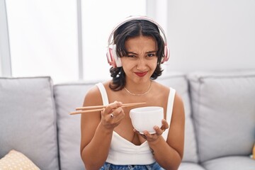 Poster - Young hispanic woman eating asian food using chopsticks clueless and confused expression. doubt concept.