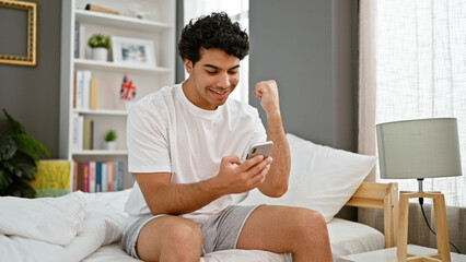 Poster - Young latin man using smartphone sitting on bed celebrating at bedroom