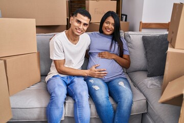 Sticker - Young hispanic couple expecting a baby sitting on the sofa at new home looking positive and happy standing and smiling with a confident smile showing teeth