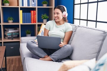 Sticker - Hispanic young woman using laptop at home wearing headphones looking away to side with smile on face, natural expression. laughing confident.