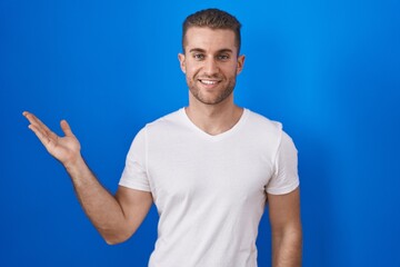 Wall Mural - Young caucasian man standing over blue background smiling cheerful presenting and pointing with palm of hand looking at the camera.