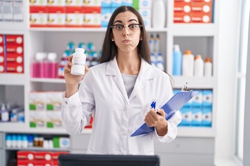 Poster - Young brunette woman working at pharmacy drugstore holding pills puffing cheeks with funny face. mouth inflated with air, catching air.