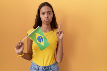 Wall Mural - Young hispanic woman holding brazil flag pointing up looking sad and upset, indicating direction with fingers, unhappy and depressed.