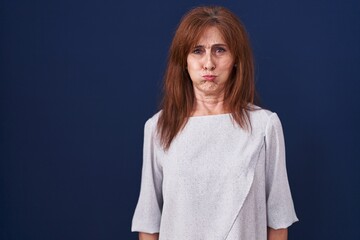 Sticker - Middle age woman standing over blue background puffing cheeks with funny face. mouth inflated with air, crazy expression.