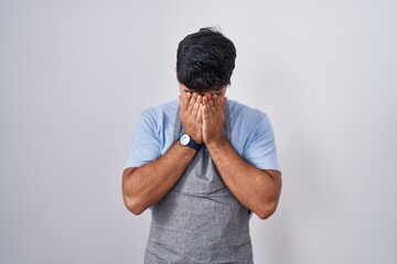 Poster - Hispanic young man wearing apron over white background with sad expression covering face with hands while crying. depression concept.