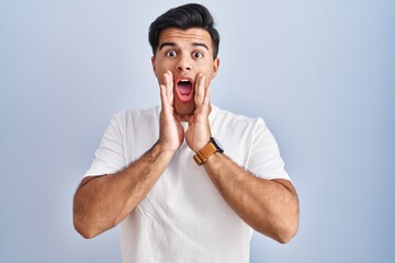 Canvas Print - Hispanic man standing over blue background afraid and shocked, surprise and amazed expression with hands on face