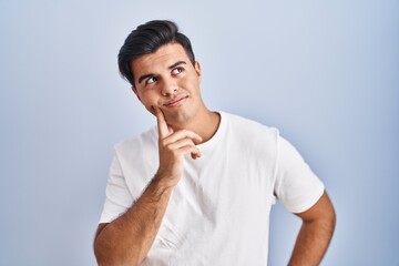 Poster - Hispanic man standing over blue background with hand on chin thinking about question, pensive expression. smiling with thoughtful face. doubt concept.