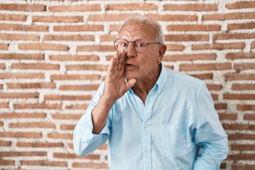 Canvas Print - Senior man with grey hair standing over bricks wall hand on mouth telling secret rumor, whispering malicious talk conversation