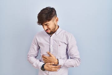 Arab man with beard standing over blue background with hand on stomach because indigestion, painful illness feeling unwell. ache concept.