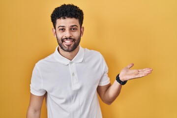 Wall Mural - Arab man standing over yellow background smiling cheerful presenting and pointing with palm of hand looking at the camera.