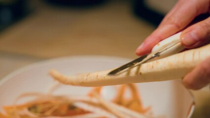 Wall Mural - Close-up of female hands peeling fresh juicy white carrot by peeler.