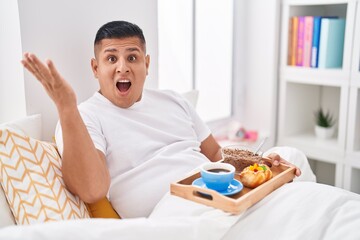 Wall Mural - Young hispanic man eating breakfast in the bed celebrating victory with happy smile and winner expression with raised hands