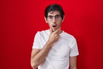 Wall Mural - Young hispanic man standing over red background looking fascinated with disbelief, surprise and amazed expression with hands on chin