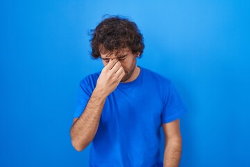 Sticker - Hispanic young man standing over blue background tired rubbing nose and eyes feeling fatigue and headache. stress and frustration concept.