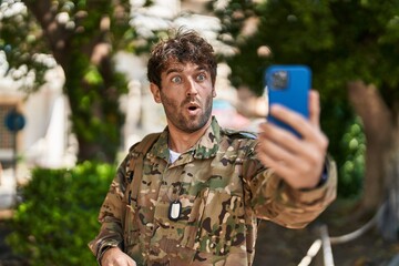 Poster - Hispanic young man wearing camouflage army uniform doing video call scared and amazed with open mouth for surprise, disbelief face