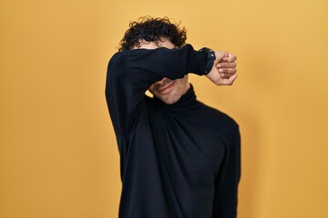 Wall Mural - Hispanic man standing over yellow background covering eyes with arm smiling cheerful and funny. blind concept.