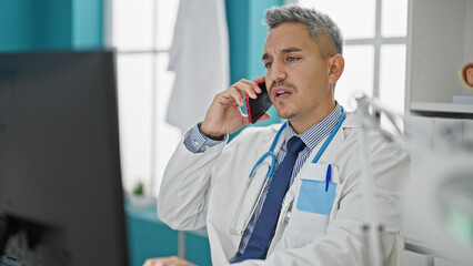 Sticker - Young hispanic man doctor using computer talking on smartphone at clinic