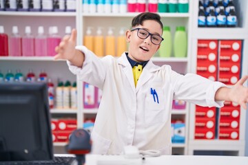 Canvas Print - Young hispanic kid working at pharmacy drugstore looking at the camera smiling with open arms for hug. cheerful expression embracing happiness.